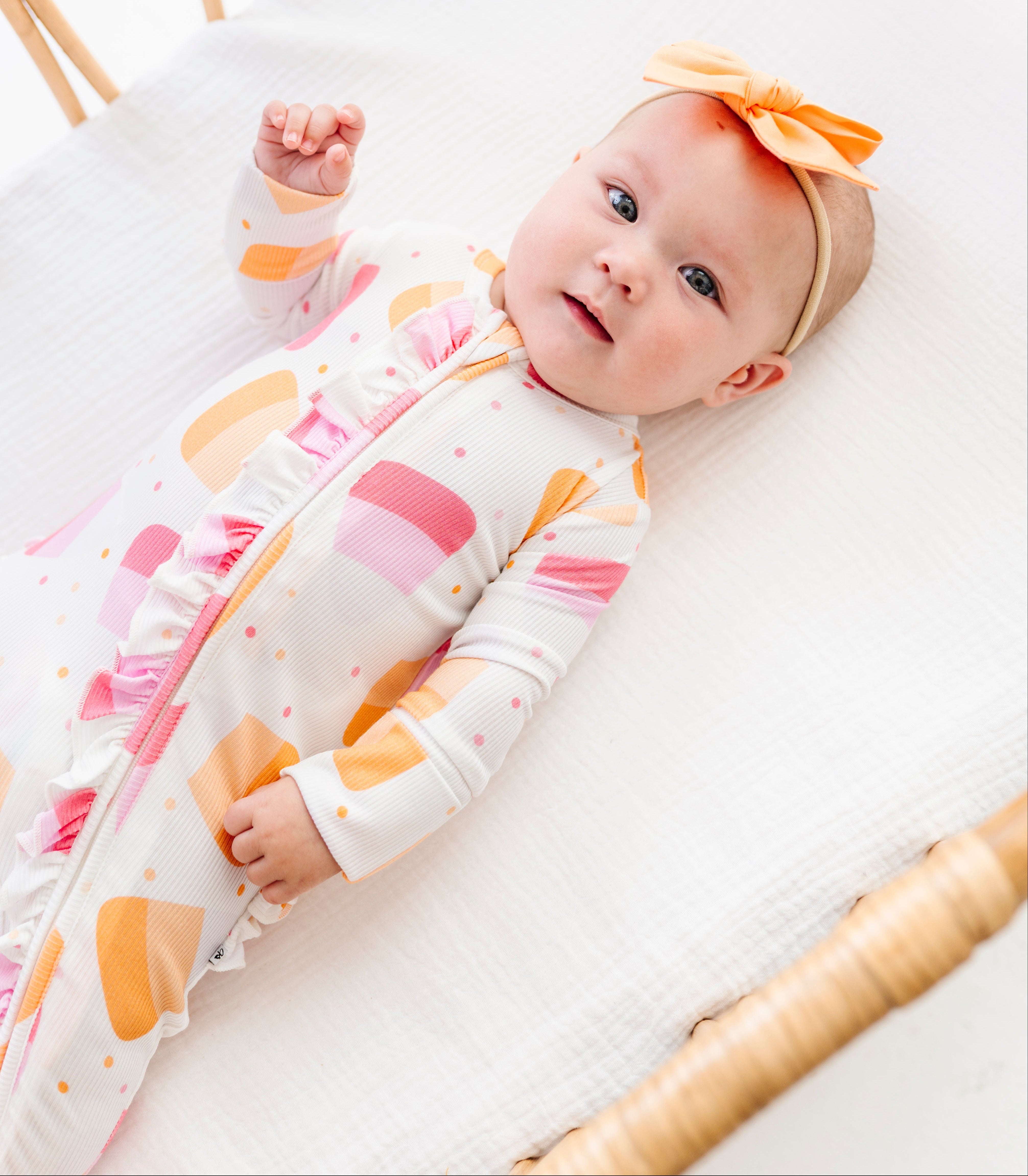 A baby lying on a white muslin mattress in a rattan bassinet, wearing a white footless sleeper with pink and orange abstract shapes and a matching orange bow headband.